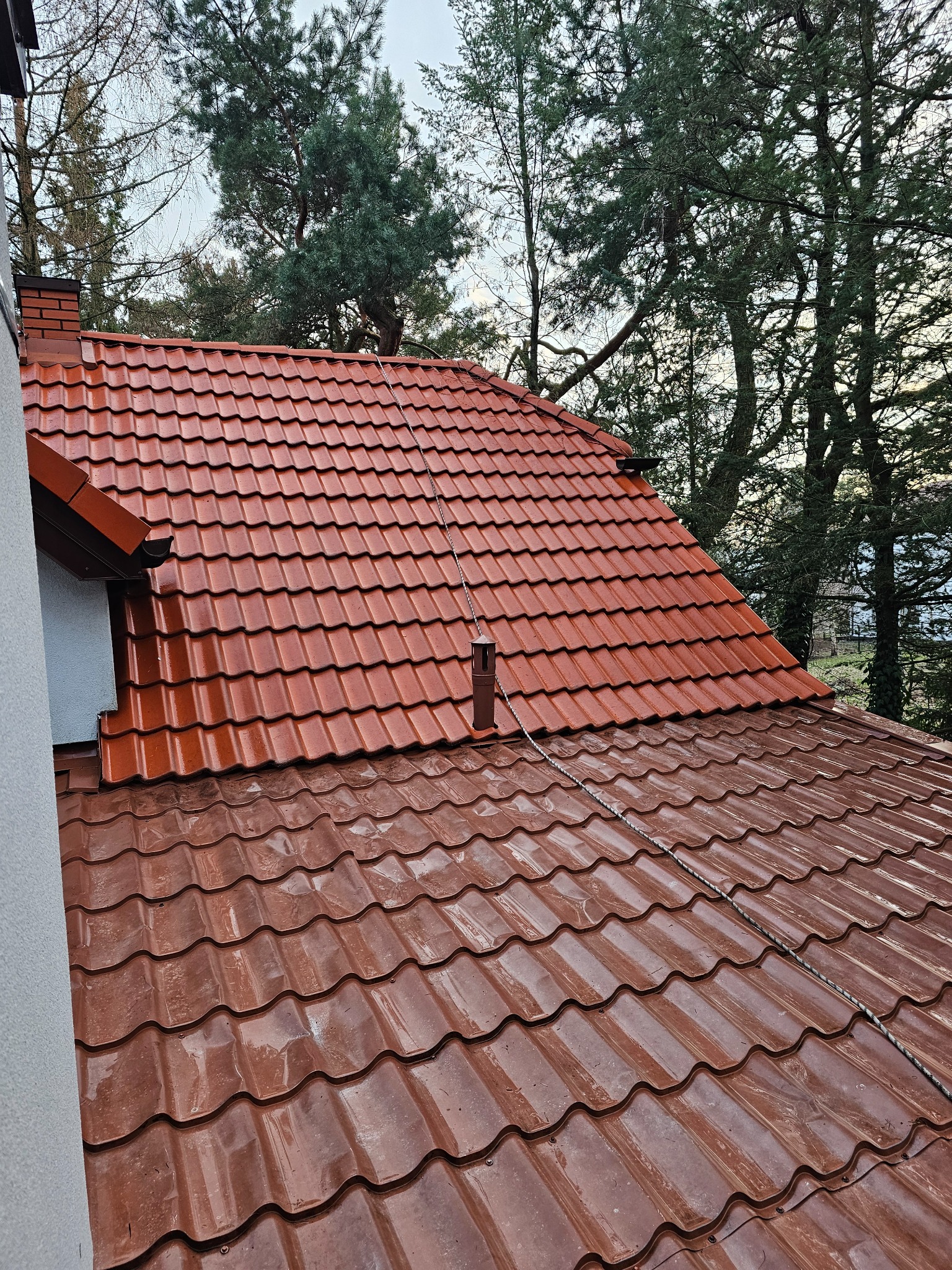 Czerwona, błyszcząca dachówka ceramiczna na ukośnym dachu domu, widok z góry, w tle drzewa i fragment komina.