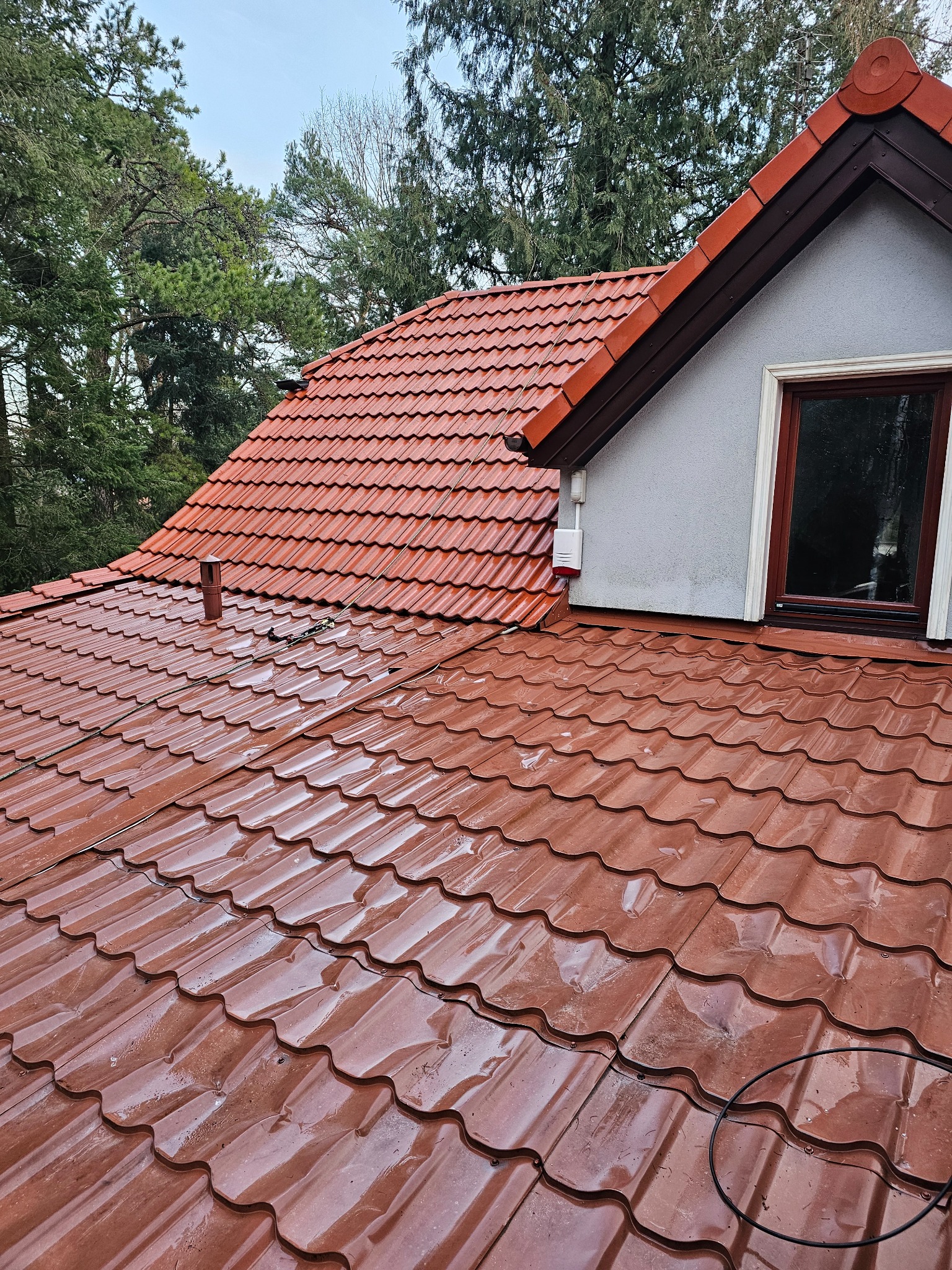 Czerwona, błyszcząca dachówka ceramiczna na skośnym dachu domu jednorodzinnego, widoczna po deszczu, z elementami systemu alarmowego, kominem i fragmentem okna poddaszowego.