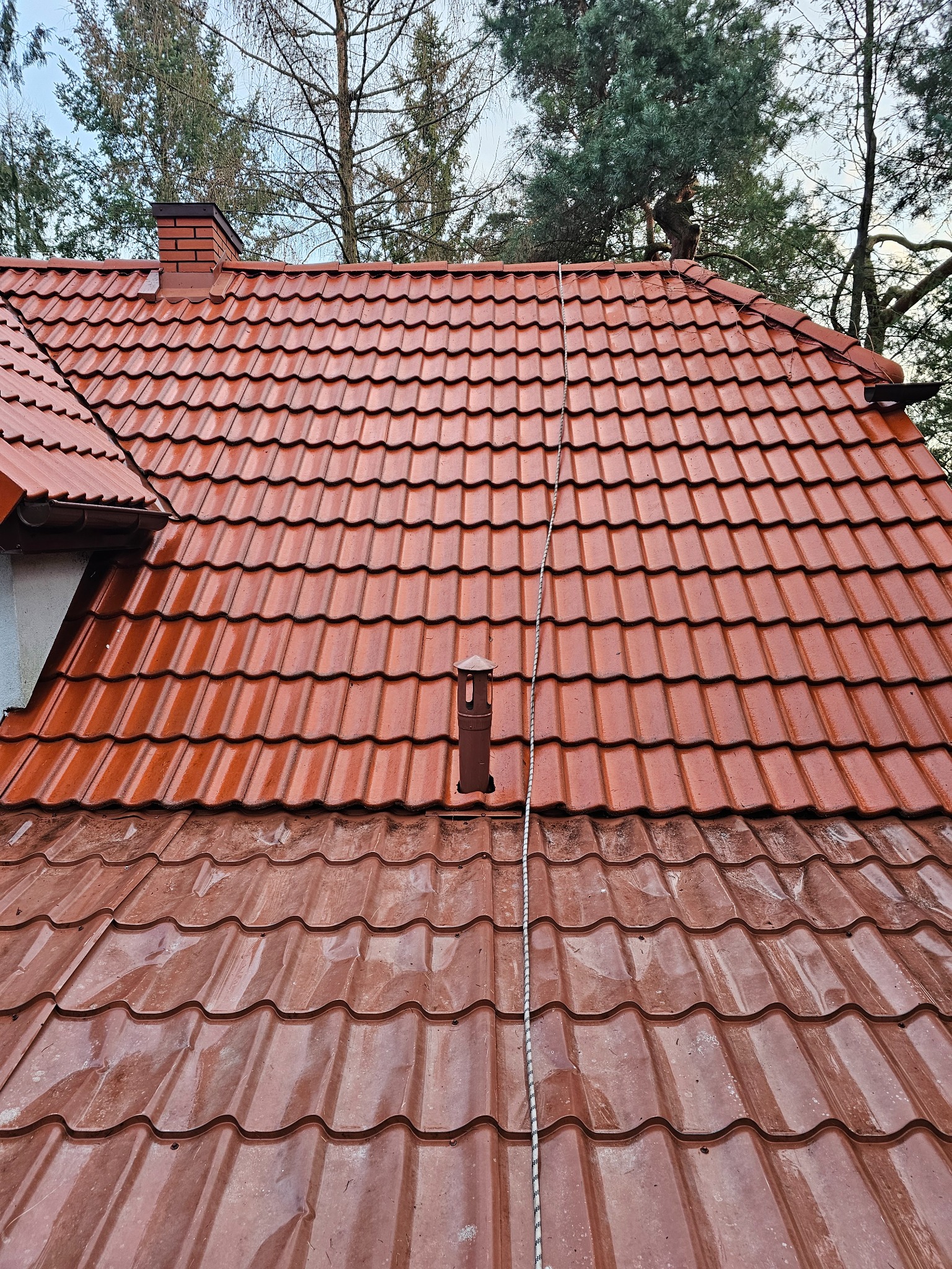 Czerwony dach z dachówki ceramicznej, częściowo wyczyszczony, z widocznym kominem i drzewami w tle.