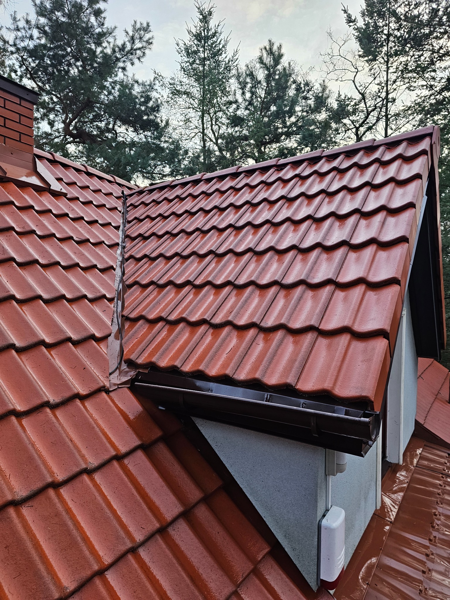 Czerwona dachówka ceramiczna na skośnym dachu domu, widok z góry, z brązowymi rynnami i kominem z cegły, na tle zielonych drzew