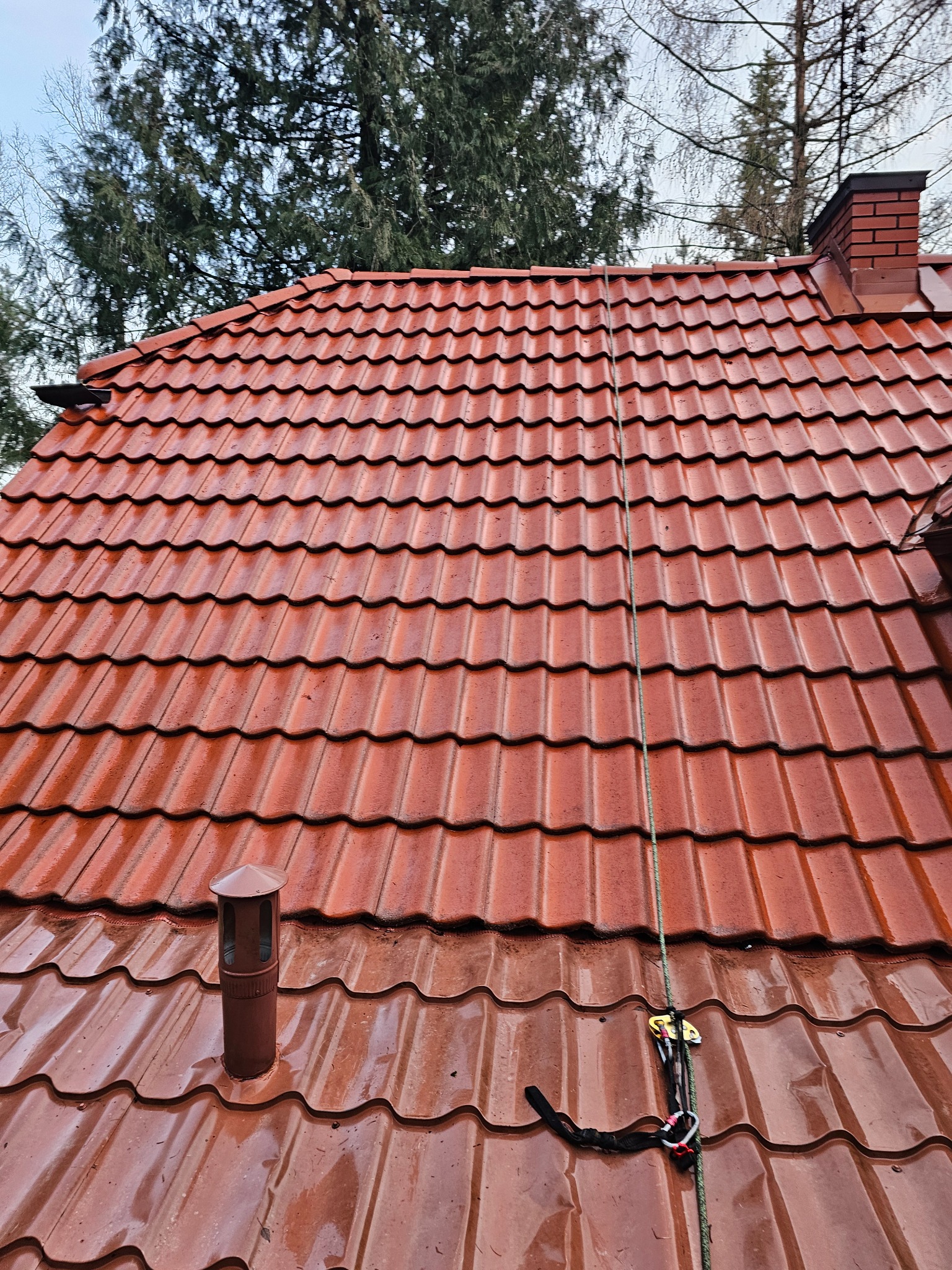 Czerwona dachówka ceramiczna, lekko wilgotna, z widocznym kominem i liną zabezpieczającą, sugerującą prace na wysokości.