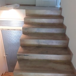 Polak Remonty - Drewniane schody wewnętrzne z subtelnymi, ciemnymi paskami antypoślizgowymi na każdym stopniu, prowadzące do białych drzwi w jasnym wnętrzu.