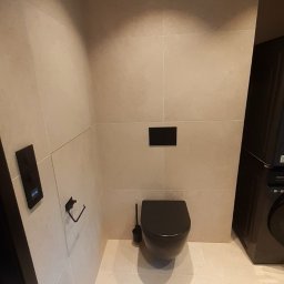 Polak Remonty - Wykończona łazienka z beżowymi płytkami, czarną toaletą i akcesoriami. Minimalistyczny design, widoczny panel sterowania i gniazdko elektryczne.