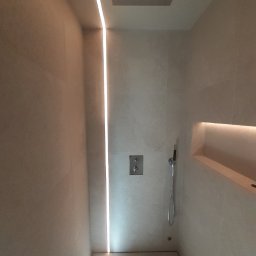 Polak Remonty - Nowoczesna kabina prysznicowa z beżowymi płytkami, liniowym odpływem i oświetleniem LED wzdłuż ściany. Minimalistyczny design z wnęką na kosmetyki.
