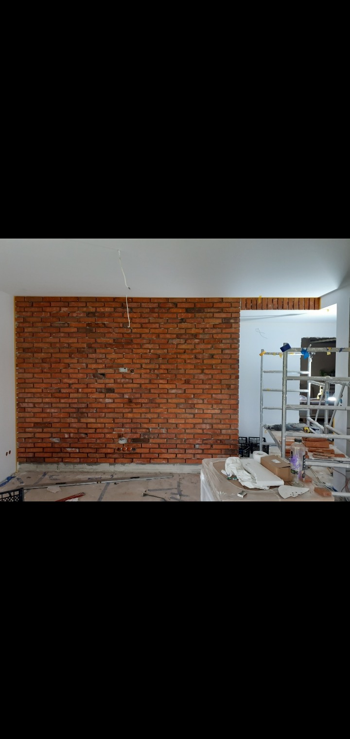 Ceglana ściana w trakcie montażu, widoczne narzędzia i rusztowanie. Częściowo wykończone wnętrze z surowym wykończeniem podłogi i widoczną instalacją elektryczną.