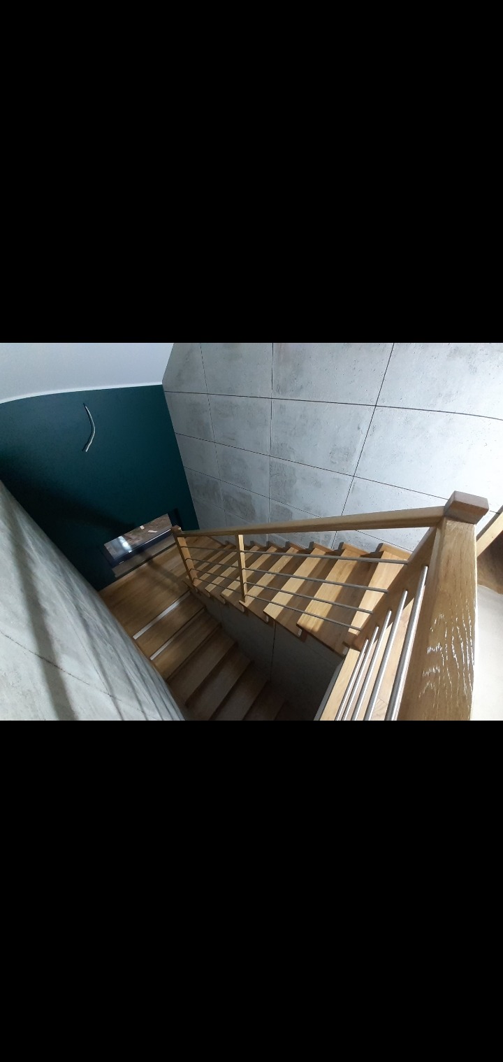 Drewniane schody z metalową balustradą, widok z góry. Szare płytki na ścianie i fragment ściany pomalowanej na ciemnozielono. Nowoczesny design.