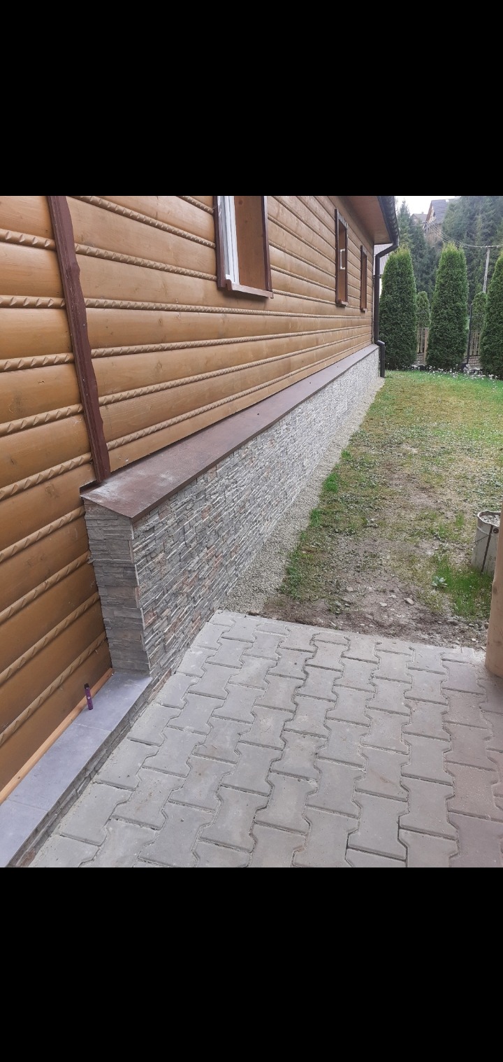 Wykończenie ściany zewnętrznej drewnianego domu kamieniem dekoracyjnym, widok z boku na cokół i chodnik z kostki brukowej. Tło: trawnik i tuje.