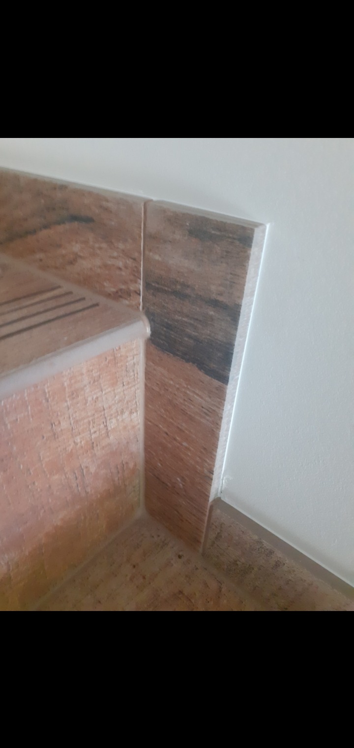 Wykończenie schodów i podłogi płytkami imitującymi drewno, z widocznymi listwami przypodłogowymi i krawędziami schodów. Detal łączenia płytek z białą ścianą.