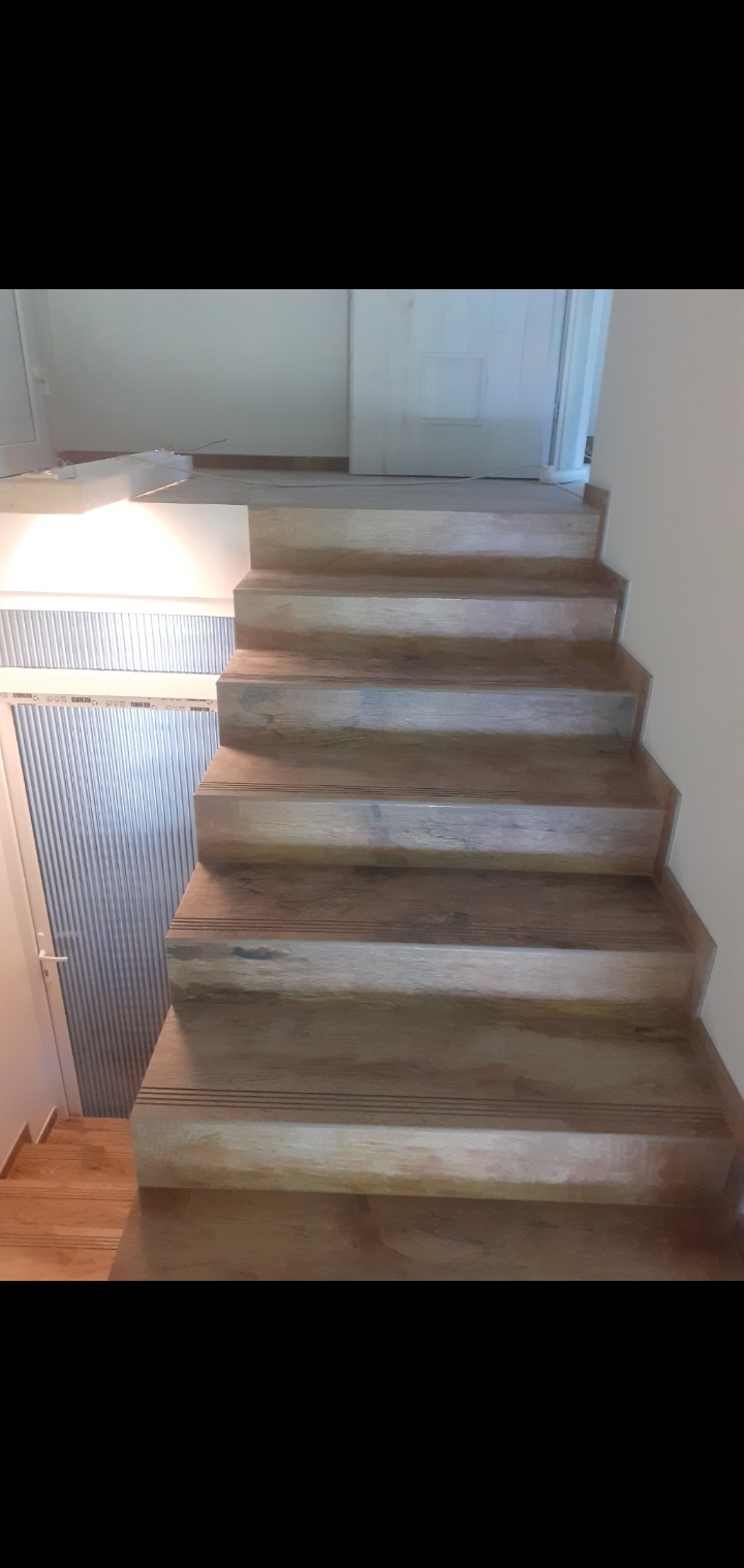 Drewniane schody wewnętrzne z subtelnymi, ciemnymi paskami antypoślizgowymi na każdym stopniu, prowadzące do białych drzwi w jasnym wnętrzu.