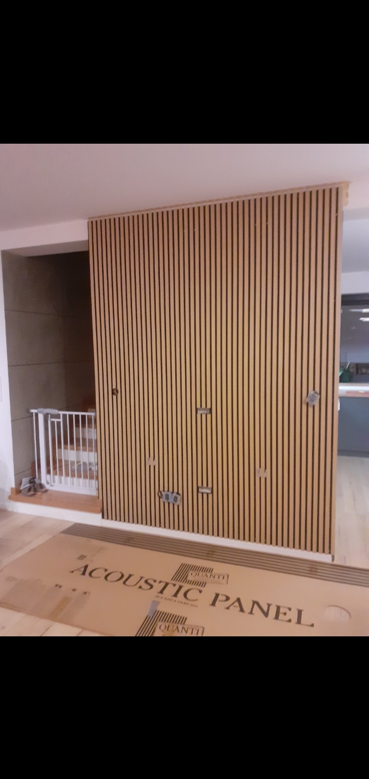 Montaż paneli akustycznych na ścianie, widoczne otwory na gniazdka elektryczne, karton z napisem 'Acoustic Panel' na podłodze, fragment schodów z barierką.