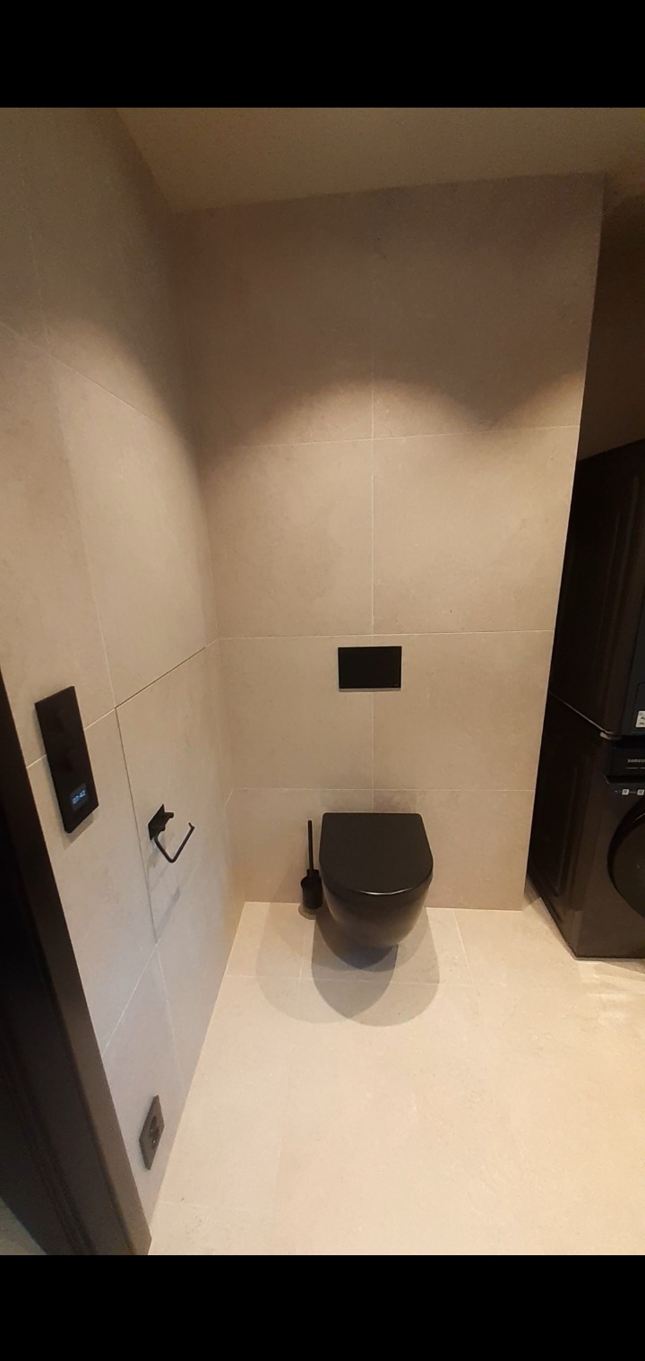 Wykończona łazienka z beżowymi płytkami, czarną toaletą i akcesoriami. Minimalistyczny design, widoczny panel sterowania i gniazdko elektryczne.