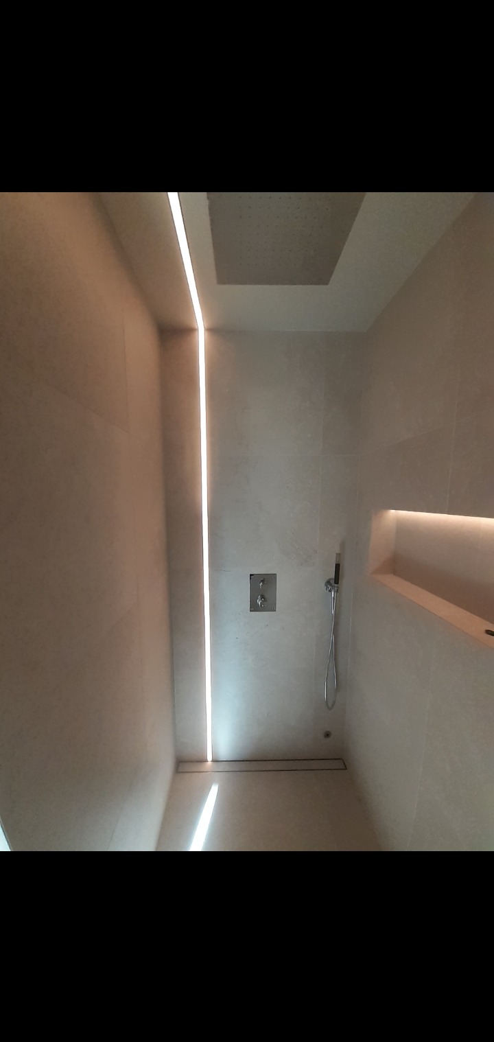 Nowoczesna kabina prysznicowa z beżowymi płytkami, liniowym odpływem i oświetleniem LED wzdłuż ściany. Minimalistyczny design z wnęką na kosmetyki.