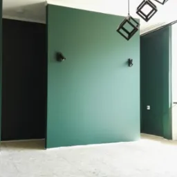 Pomalowane na zielono ściany z zamontowanymi reflektorami, sufit biały, podłoga z płyt OSB, wiszące lampy w kształcie czarnych sześcianów.