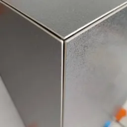 Zbliżenie na narożnik wykończony szarymi płytkami o delikatnej strukturze, widoczne krzyżyki dystansowe w kolorze pomarańczowym i niebieskim.