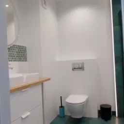 Nowoczesna łazienka z podwieszaną toaletą, białą umywalką nablatową na drewnianym blacie i zieloną podłogą z efektem betonu.