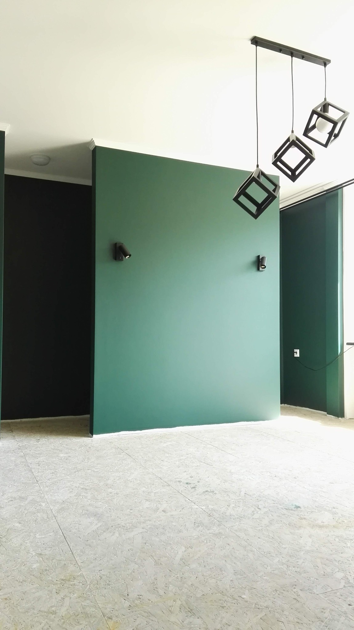 Pomalowane na zielono ściany z zamontowanymi reflektorami, sufit biały, podłoga z płyt OSB, wiszące lampy w kształcie czarnych sześcianów.