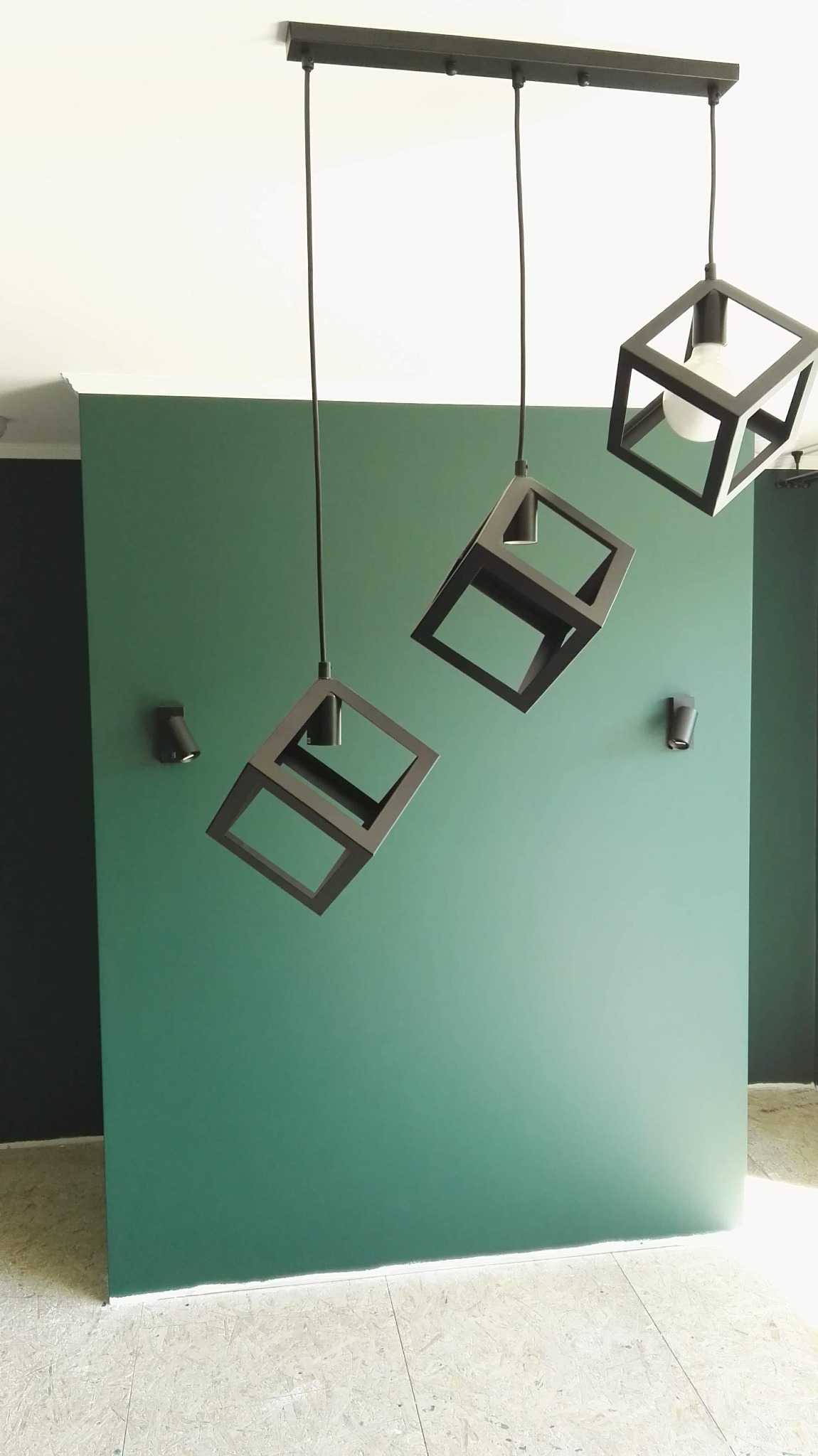 Trzy czarne, geometryczne lampy wiszące w kształcie sześcianów na tle zielonej ściany z dwoma małymi reflektorami punktowymi.