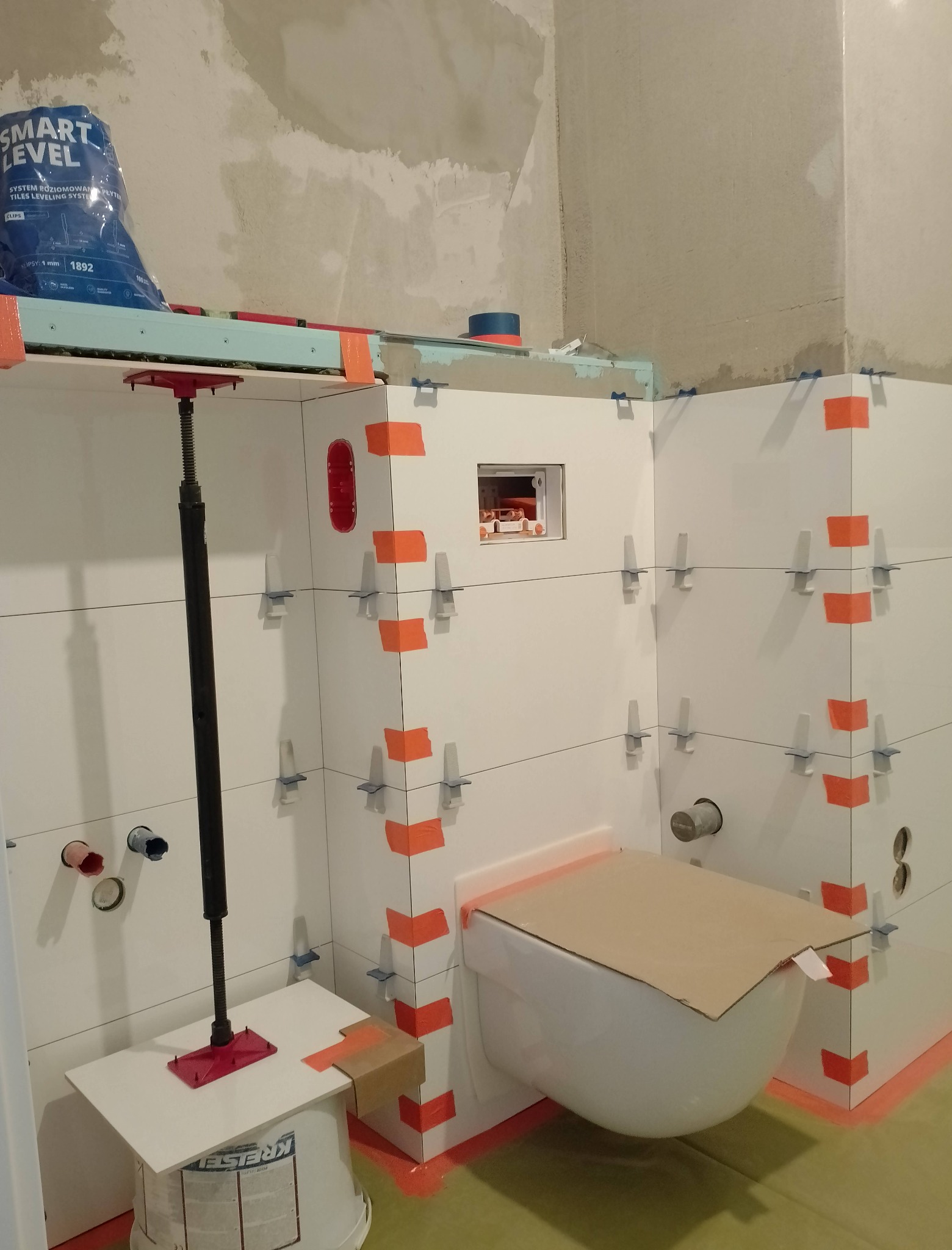 Białe płytki ścienne w trakcie montażu, z użyciem pomarańczowej taśmy i klipsów poziomujących, widoczna podwieszana toaleta zabezpieczona kartonem, narzędzia i worek z zaprawą w tle.