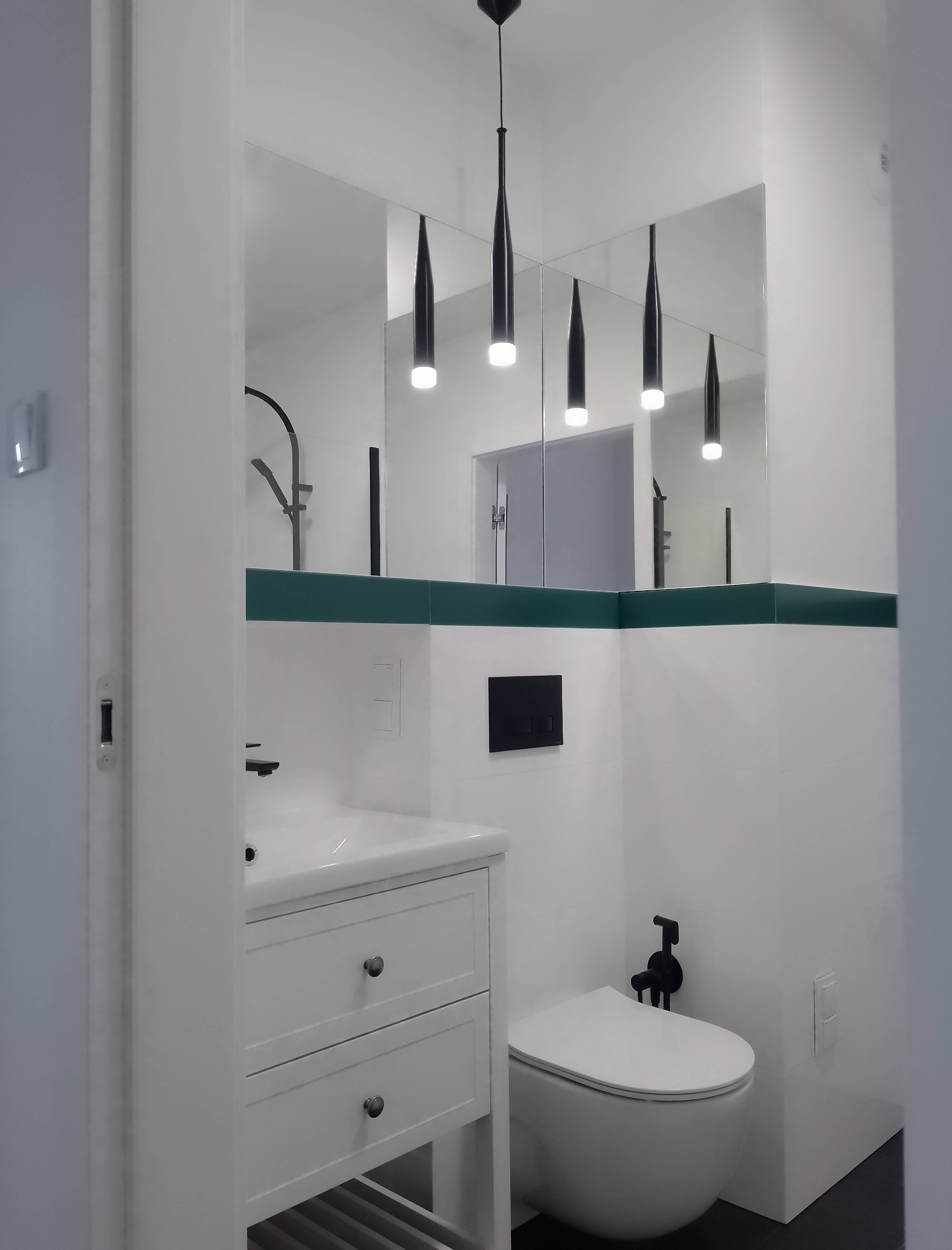 Nowoczesna łazienka w bieli z akcentami czerni i butelkowej zieleni; wisząca toaleta z czarną armaturą, biała szafka pod umywalkę z dwiema szufladami, lustro z zielonym pasem płytek i czarne...