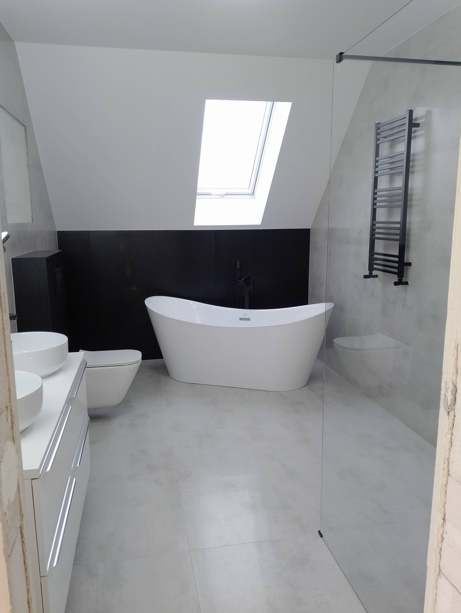 Nowoczesna łazienka na poddaszu z białą wolnostojącą wanną, umywalką nablatową, toaletą podwieszaną i czarnym grzejnikiem drabinkowym na tle szarej ściany i czarnej zabudowy.