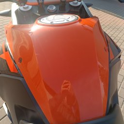 AUTO-DETAILING KAMIL KNOP - Pomarańczowy bak motocykla KTM z widocznym w odbiciu zarysem dłoni. Detale takie jak wlew paliwa i czarne elementy nadwozia. Kostka brukowa w tle.