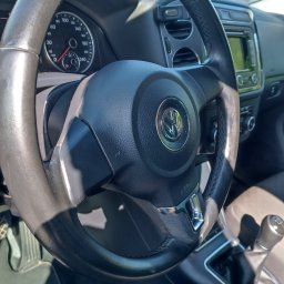 Auto-Detailing Kamil Knop - Czarne koło kierownicy z logo VW wewnątrz pojazdu, widoczny kokpit i fragment fotela pasażera, światło słoneczne.