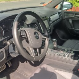 Auto-Detailing Kamil Knop - Wnętrze samochodu Volkswagen Touareg z czarną skórzaną tapicerką, widok na deskę rozdzielczą, kierownicę z logo VW i automatyczną skrzynię biegów. Jasne oświetlenie.