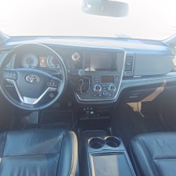 Auto-Detailing Kamil Knop - Wnętrze samochodu Toyota Sienna: czarna skórzana tapicerka foteli, deska rozdzielcza z ekranem dotykowym, kierownica z logo Toyota, jasne światło wpadające przez przednią szybę.