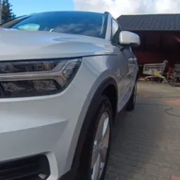 Biały samochód po usłudze mycia, zaparkowany na kostce brukowej w słoneczny dzień. Widoczny fragment karoserii, koło i reflektor. W tle budynek z ciemnym dachem.