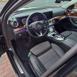 Auto-Detailing Kamil Knop - Wnętrze czarnego Mercedesa E-Klasy z drewnianymi akcentami, widok z otwartymi drzwiami na deskę rozdzielczą, kierownicę i fotele, z widocznym logo marki.