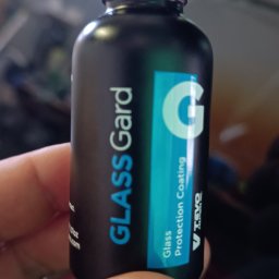 AUTO-DETAILING KAMIL KNOP - Czarna butelka z etykietą 'Glass Gard Glass Protection Coating' trzymana w dłoni, z widocznym fioletowym płynem wewnątrz. Błyszcząca powierzchnia sugeruje ochronę szkła.