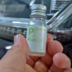 AUTO-DETAILING KAMIL KNOP - Dłoń trzyma buteleczkę środka PlusGard do pielęgnacji plastiku, gumy i skóry na tle fragmentu karoserii samochodowej. Srebrna zakrętka.