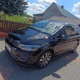 AUTO-DETAILING KAMIL KNOP - Lśniący, czarny minivan na brukowanym podjeździe w słoneczny dzień. Karoseria odbija błękitne niebo z chmurami. Widoczny dom i ogrodzenie w tle.