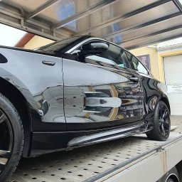AUTO-DETAILING KAMIL KNOP - Błyszczący, czarny samochód na lawecie pod zadaszeniem myjni, odbijający otoczenie w lakierze. Widoczny fragment opony i karoserii.