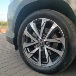 Felga aluminiowa z logo Toyota na oponie Dunlop, widoczna na tle fragmentu karoserii auta w odcieniach zieleni i szarości, zaparkowanego na szarej kostce brukowej.
