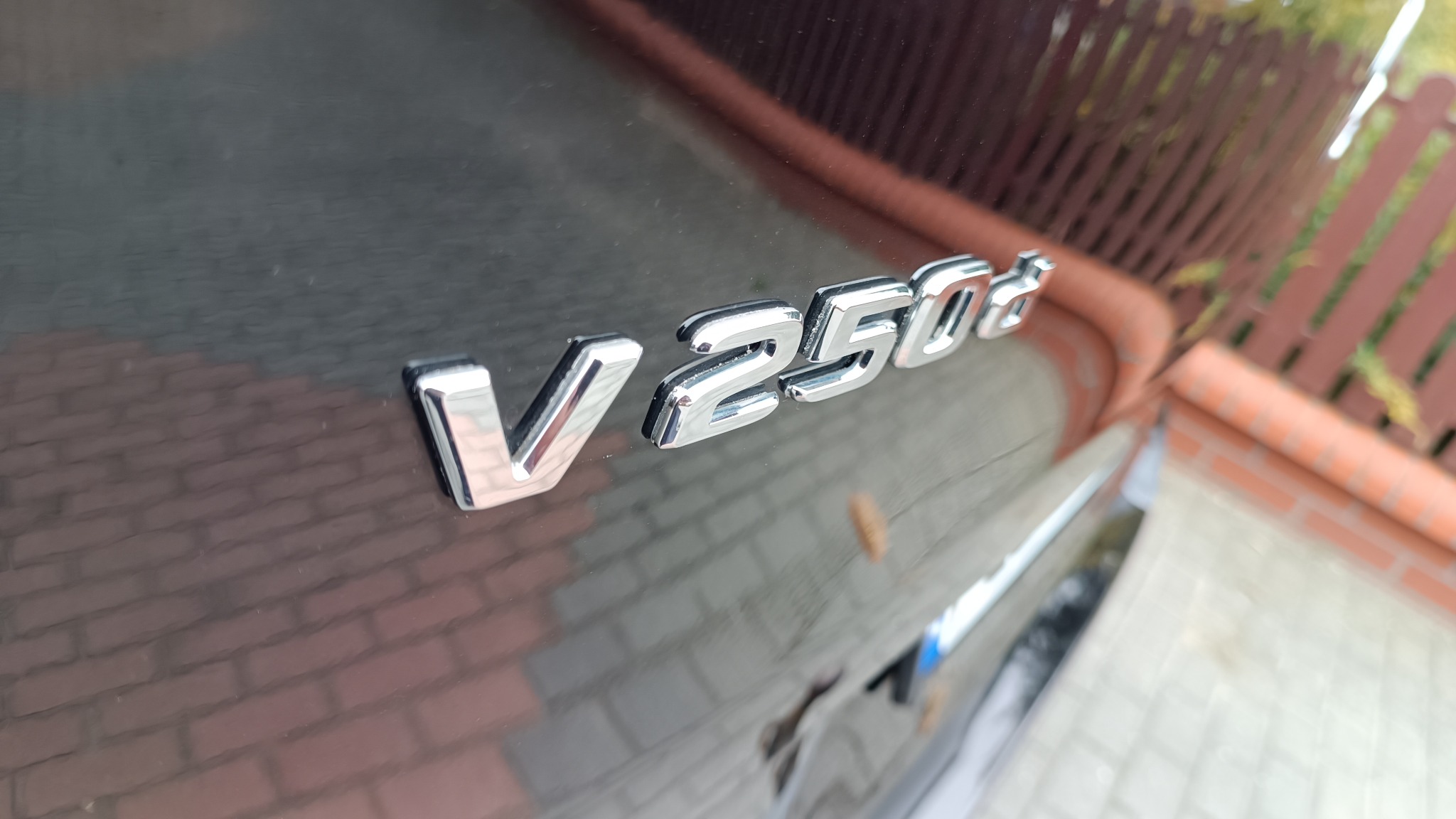 Błyszczący chromowany napis 'V 250 d' na ciemnym lakierze samochodu, widoczne detale karoserii i fragment otoczenia.