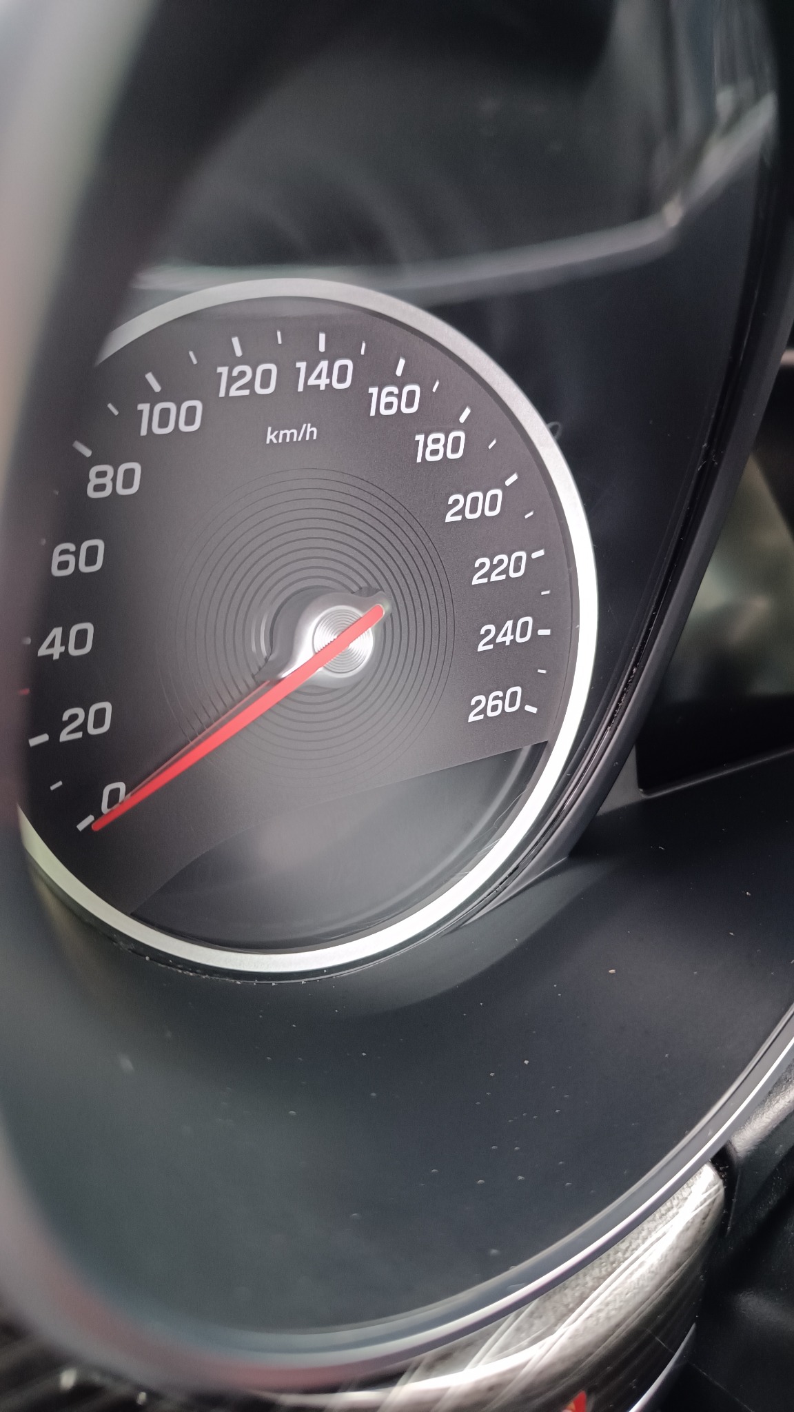 Czarny samochodowy prędkościomierz z czerwoną wskazówką, wskazujący prędkość około 20 km/h. Widoczne drobne zabrudzenia na desce rozdzielczej.