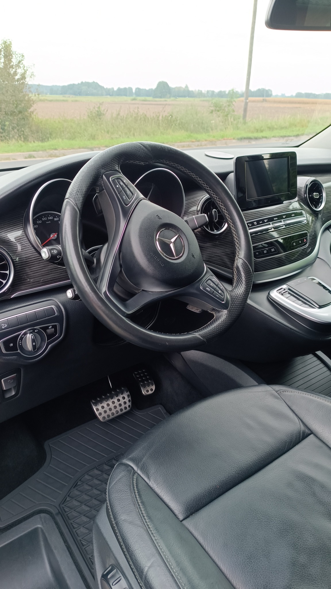 Wnętrze samochodu Mercedes V-klasa: czarna skórzana tapicerka fotela kierowcy, kierownica z logo, deska rozdzielcza z elementami drewna i metalowe nakładki na pedały.