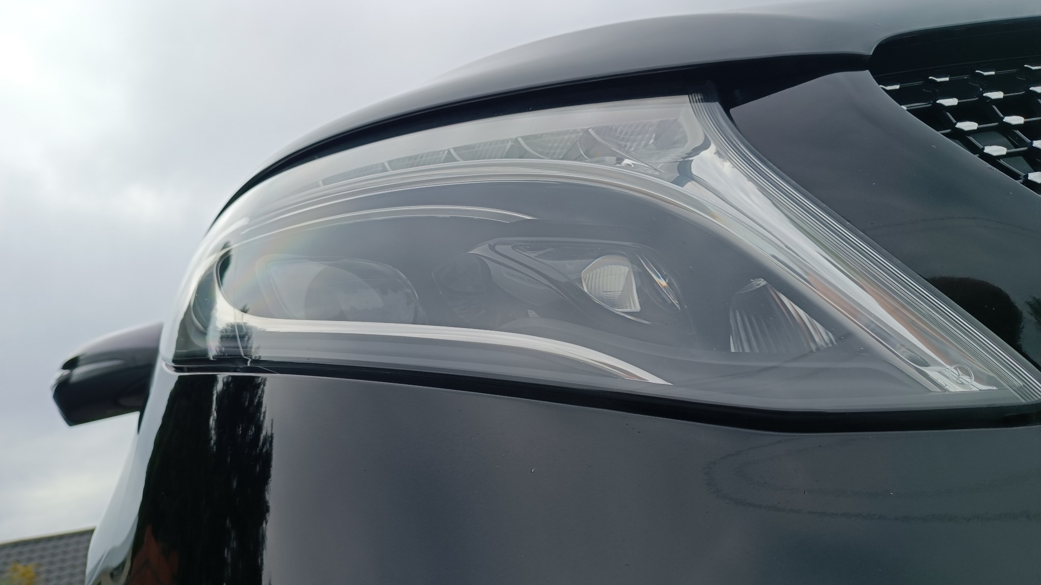 Detal czarnego samochodu: reflektor z wyraźnym odbiciem nieba. Widoczna krata wlotu powietrza i fragment karoserii.