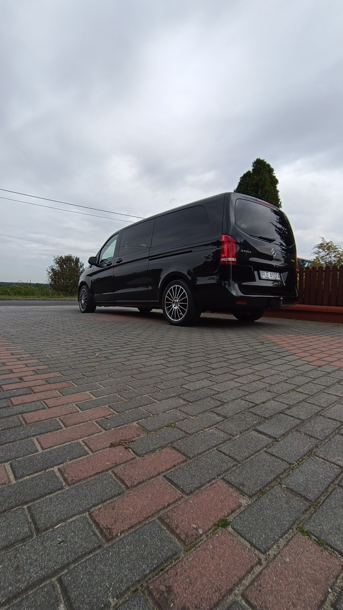 Czarny, lśniący bus Mercedes V250d z ciemnymi szybami i aluminiowymi felgami zaparkowany na kostce brukowej. Widok z tyłu, pochmurne niebo.