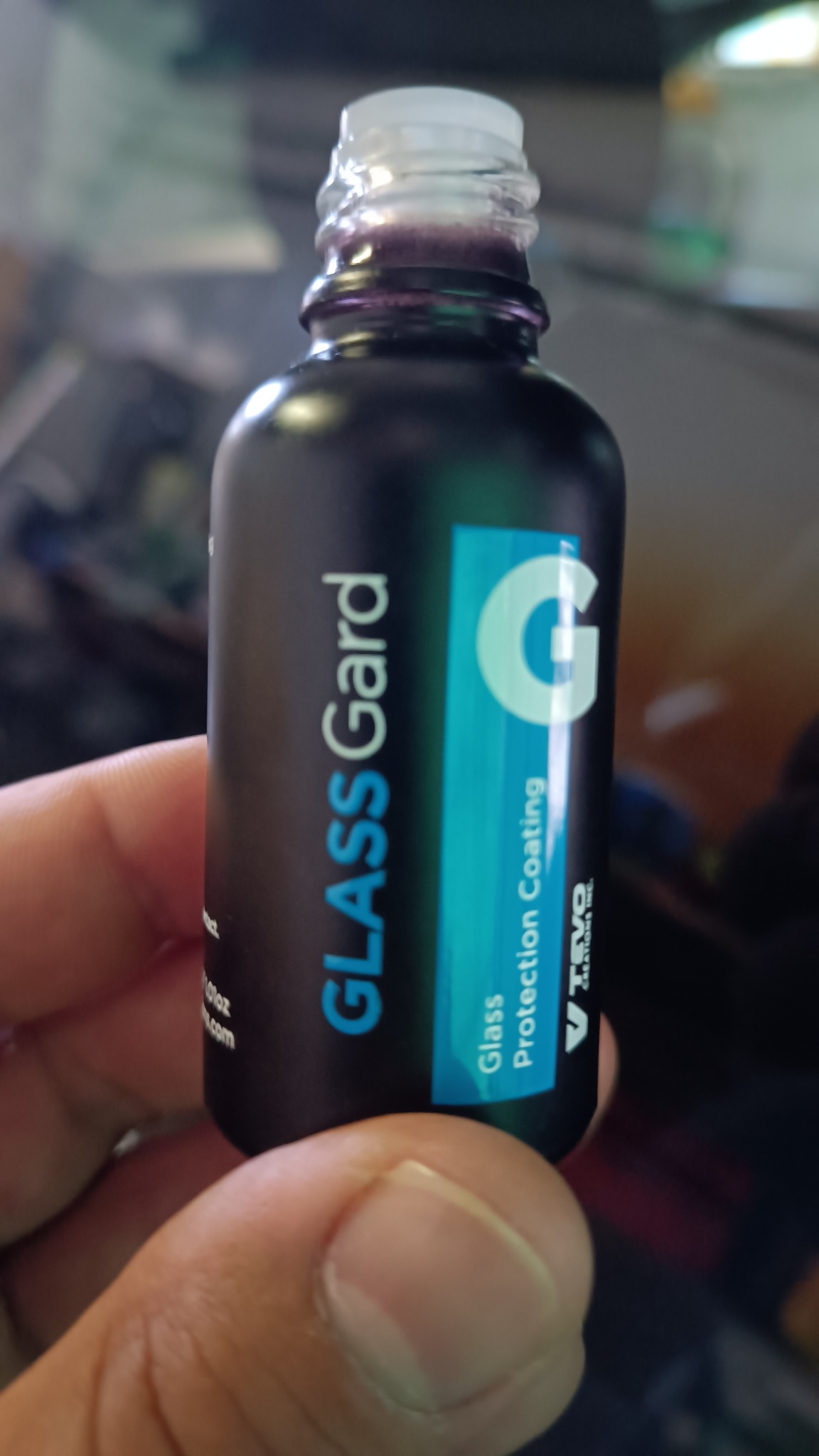Czarna butelka z etykietą 'Glass Gard Glass Protection Coating' trzymana w dłoni, z widocznym fioletowym płynem wewnątrz. Błyszcząca powierzchnia sugeruje ochronę szkła.