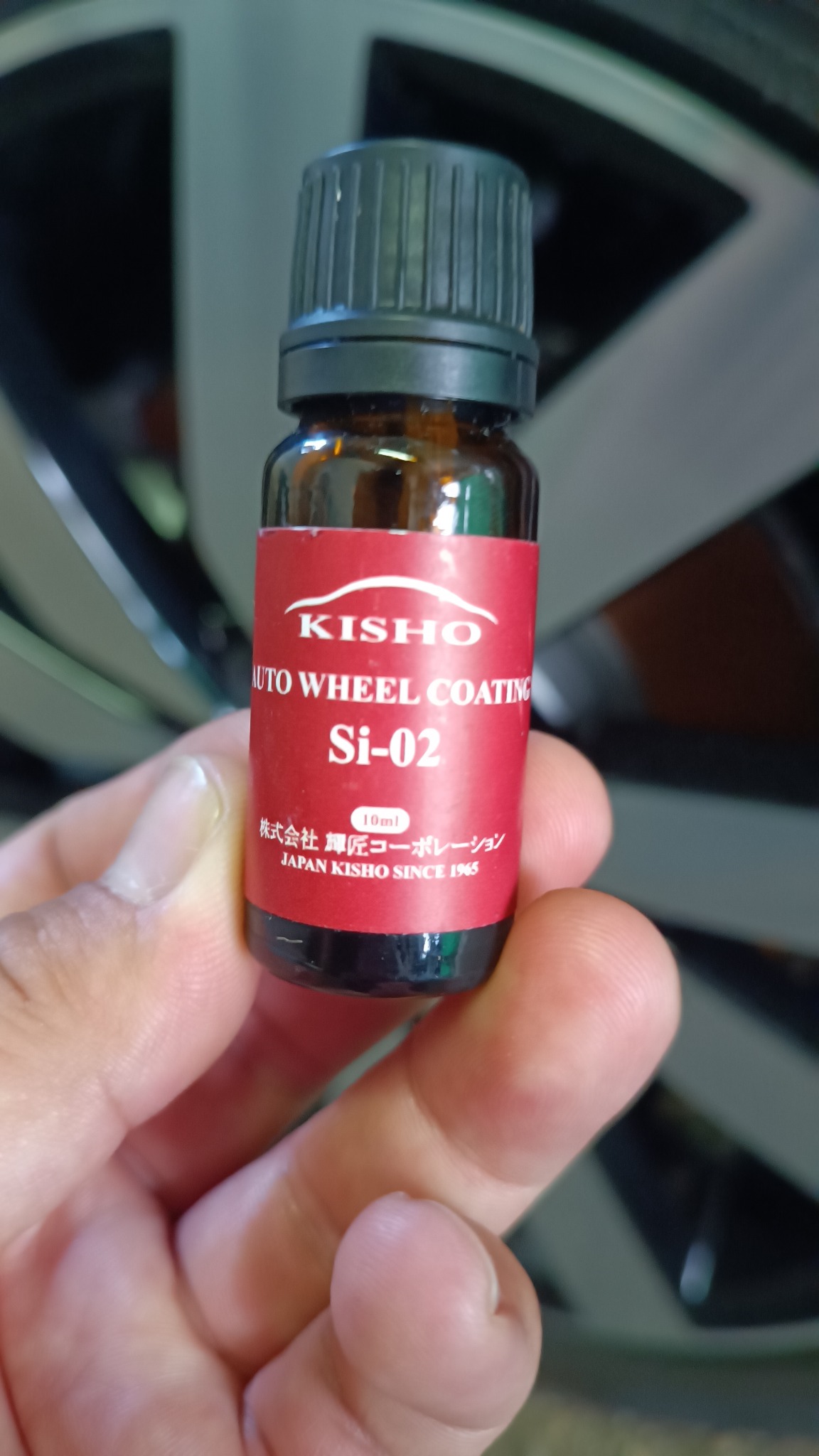 Dłoń trzyma buteleczkę KISHO Auto Wheel Coating Si-02 na tle felgi samochodowej. Produkt do konserwacji i pielęgnacji felg, zapewniający ochronę i połysk.