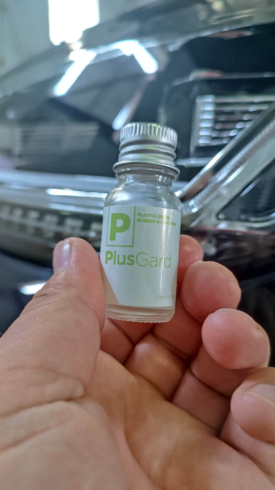 Dłoń trzyma buteleczkę środka PlusGard do pielęgnacji plastiku, gumy i skóry na tle fragmentu karoserii samochodowej. Srebrna zakrętka.