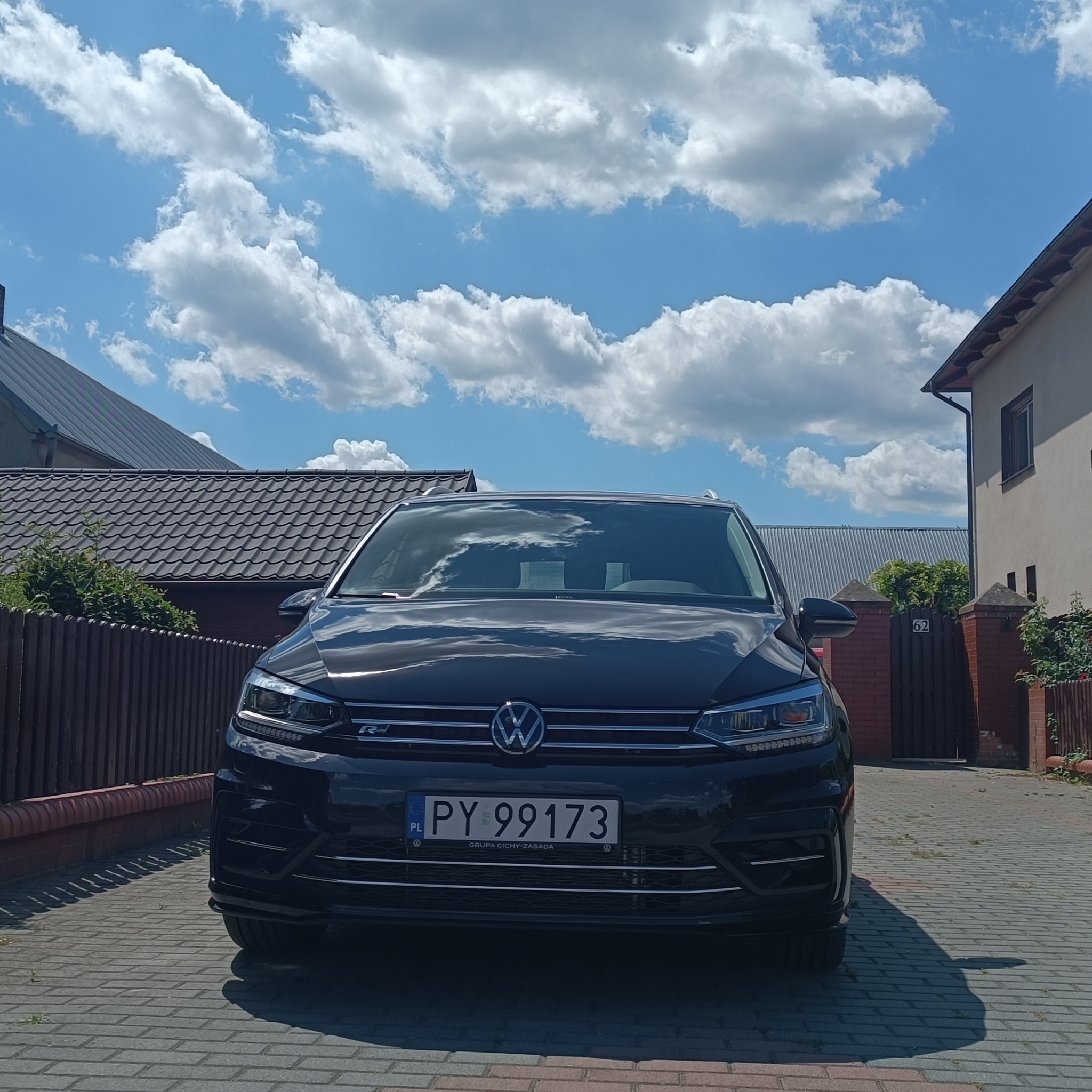 Czarny Volkswagen Touran R-Line z polską tablicą rejestracyjną PY 99173 na tle słonecznego nieba z chmurami i fragmentów budynków.