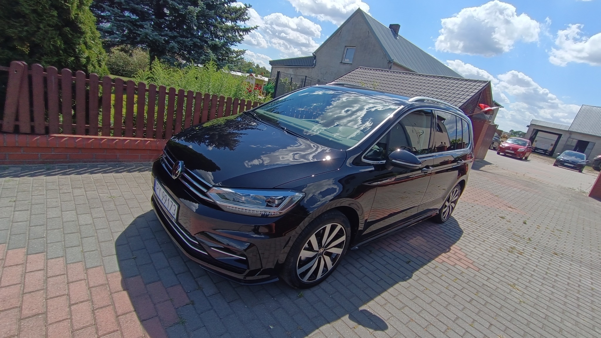 Lśniący, czarny minivan na brukowanym podjeździe w słoneczny dzień. Karoseria odbija błękitne niebo z chmurami. Widoczny dom i ogrodzenie w tle.