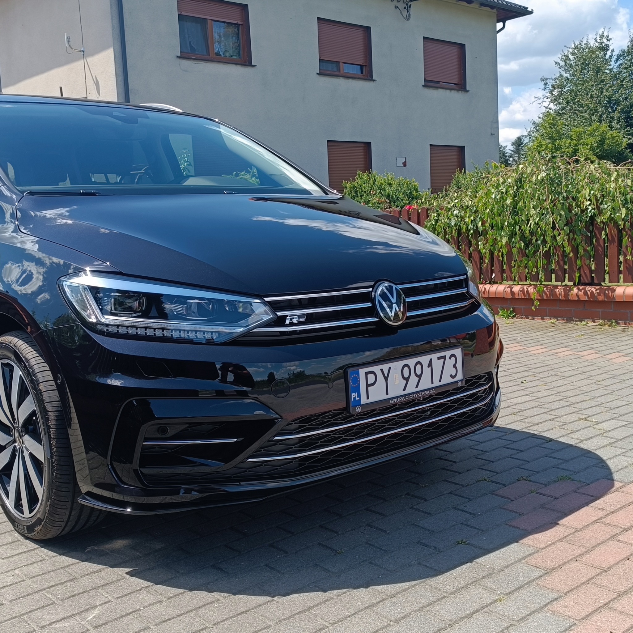 Czarny samochód Volkswagen R-Line z widoczną tablicą rejestracyjną PY 99173, zaparkowany przed budynkiem z brązowymi roletami i ogrodzeniem.