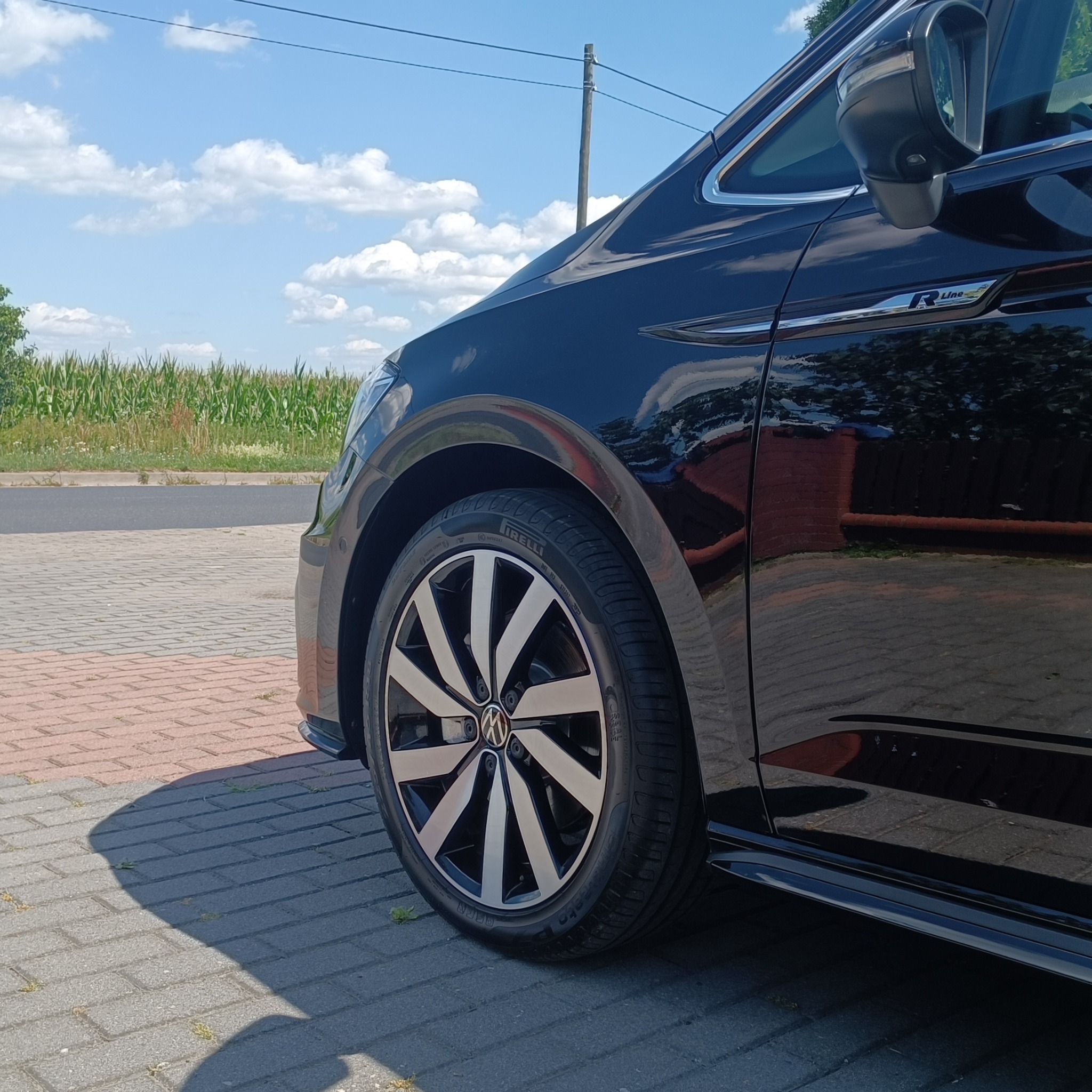 Błyszczące, czarne auto R-Line z felgami aluminiowymi na tle błękitnego nieba i pola kukurydzy. Widoczny fragment karoserii, koło i opona.