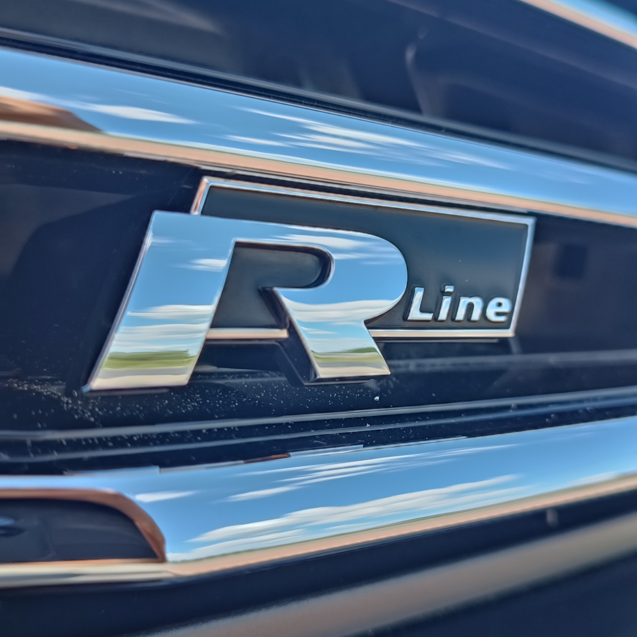 Błyszczące logo 'R Line' na czarnym grillu samochodu, odbijające niebo. Chromowane detale i precyzyjne wykonanie.