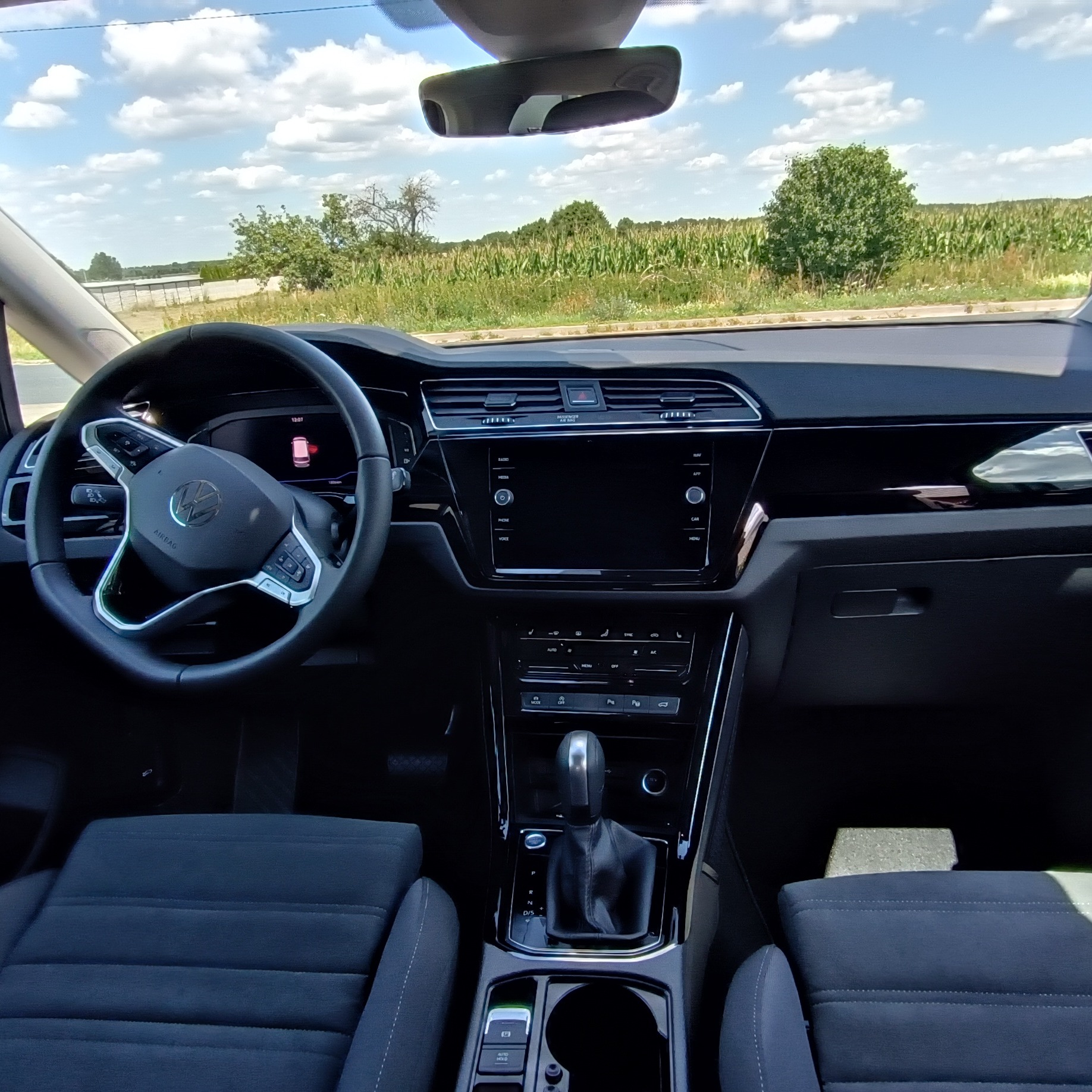 Wnętrze samochodu Volkswagen z widokiem na deskę rozdzielczą, kierownicę i przednią szybę z krajobrazem. Dominują ciemne kolory tapicerki.