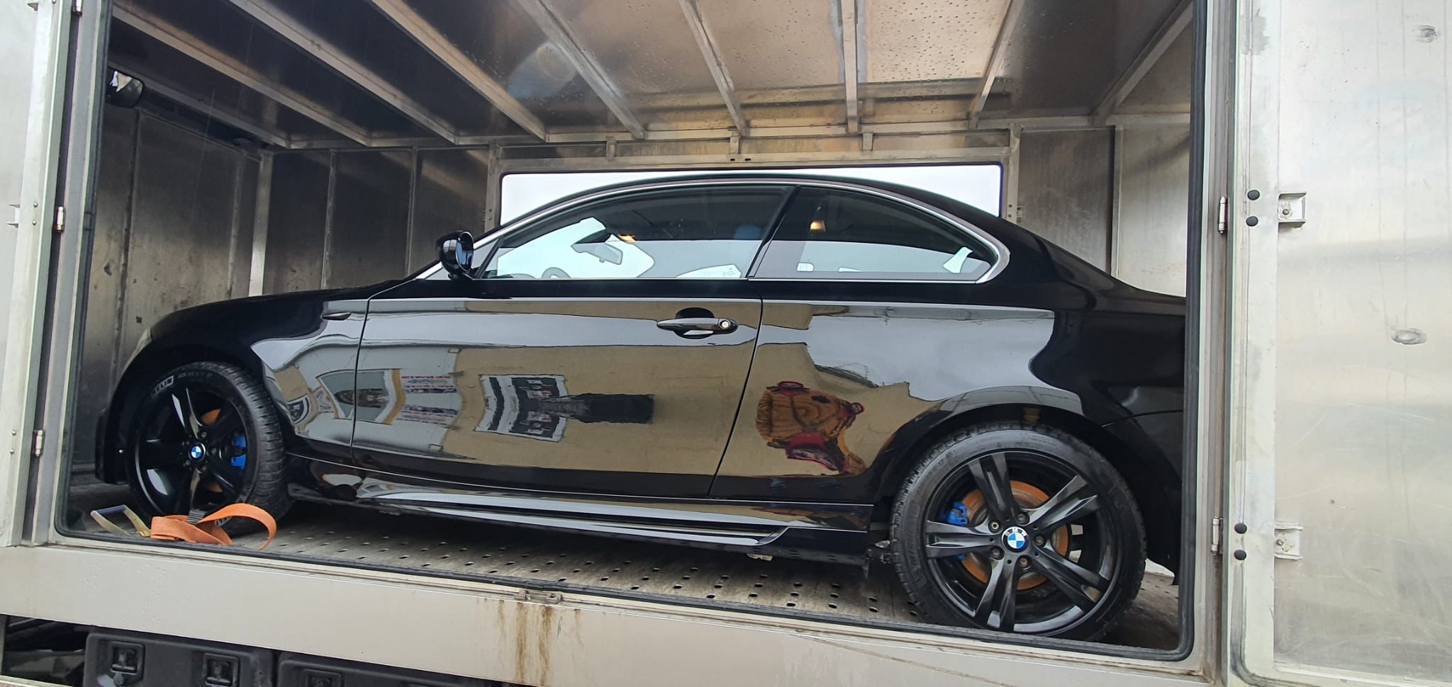 Błyszczące, czarne BMW w przestrzeni ładunkowej, widok z boku. Felgi z niebieskimi zaciskami hamulcowymi, pomarańczowy pas transportowy. Metalowe wnętrze pojazdu.